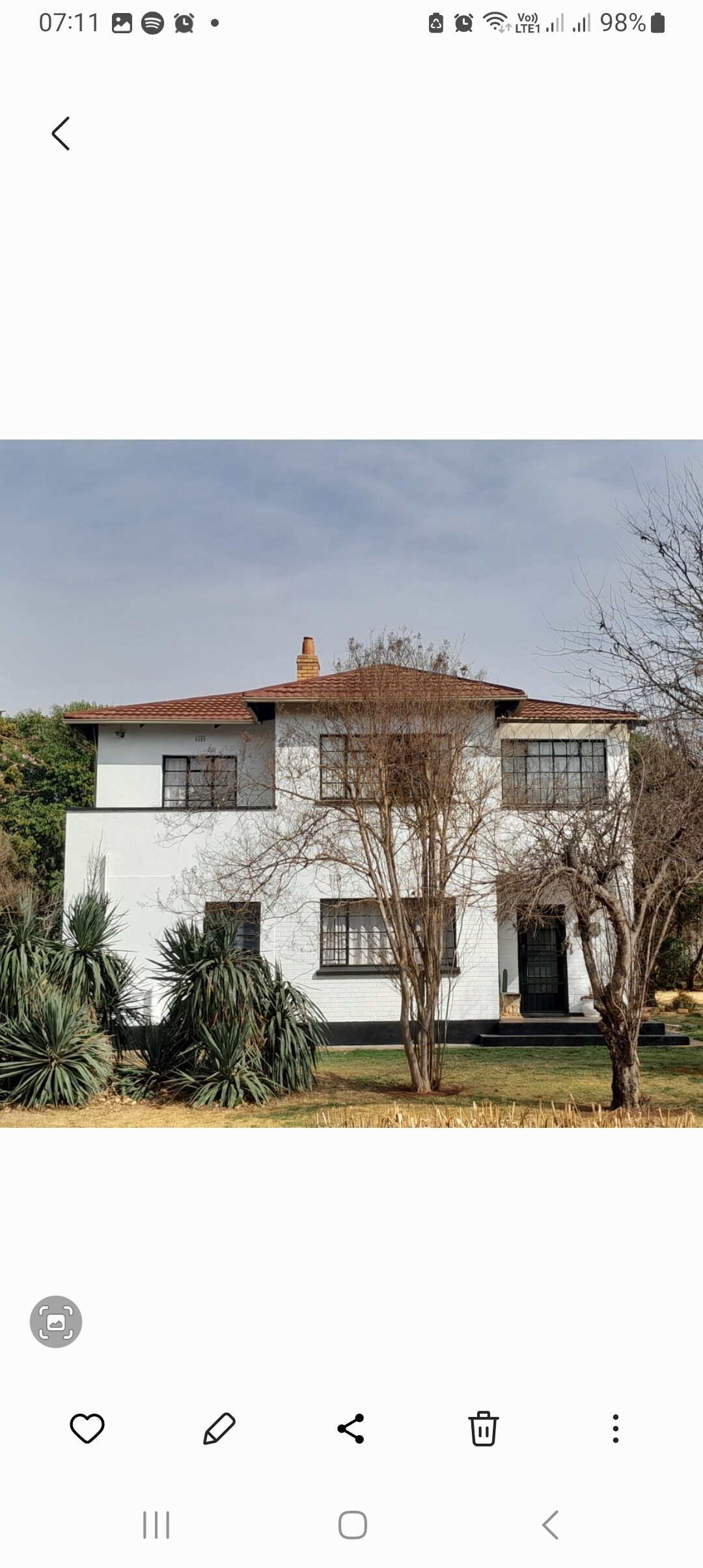 A semi-detached, two-story building is shown with a white exterior and dark window frames. The structure is set in a landscaped garden featuring various shrubs and plants. A clear sky is visible above, enhancing the serene ambiance of the setting.