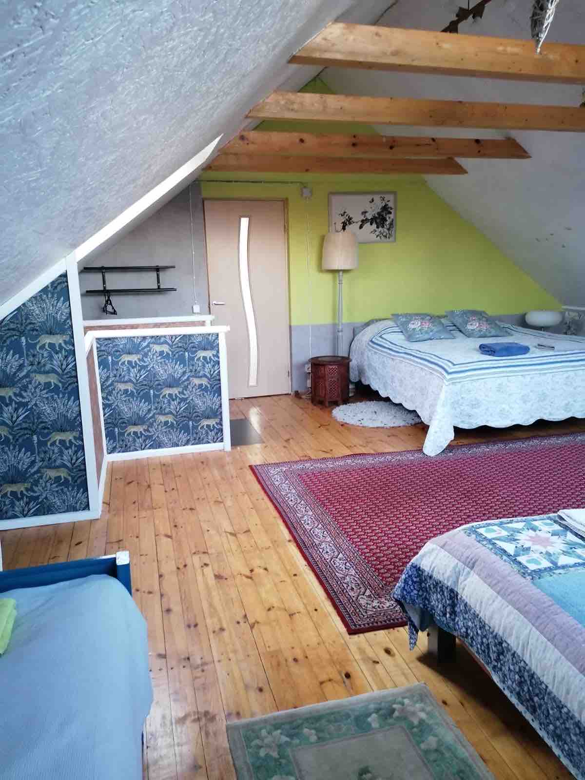 A spacious bedroom features sloped ceilings and wooden floors. Two beds are arranged in the room, with colorful quilts on each. A decorative area rug adds warmth, while bright green and blue walls bring a vibrant touch. Natural light filters in from a nearby window.