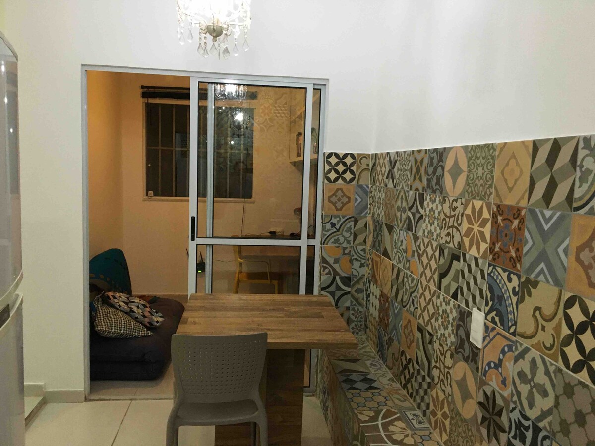 A compact dining area features a wooden table paired with a textured plastic chair. A vibrant tiled wall, adorned with various geometric patterns, adds character to the space. A small window allows natural light to fill the room, creating a cozy atmosphere.