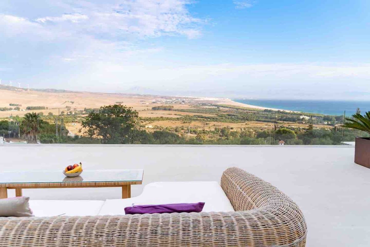 An outdoor seating area is visible, featuring a curved wicker couch with white cushions. A glass table holds a bowl of fruit. The expansive view includes rolling hills and the sea, with the sky showcasing a mix of clouds and sunlight.