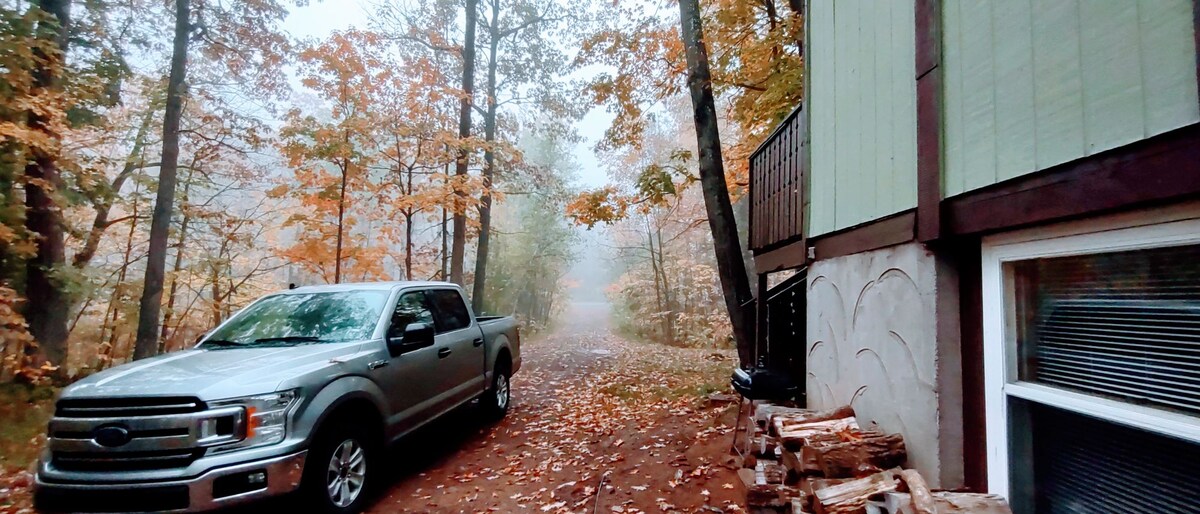 A silver pickup truck is parked along a gravel path, surrounded by a vibrant autumn landscape. Colorful leaves cover the ground, while misty air creates a serene atmosphere among the trees lining the route. The house's exterior features a muted green and brown color scheme.