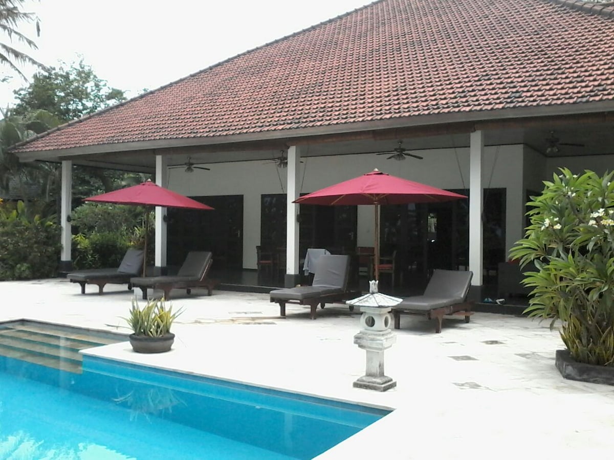 An inviting outdoor area features a swimming pool surrounded by lounge chairs with red umbrellas for shade. A spacious terrace is accessible from the villa, showcasing a comfortable seating arrangement and lush tropical vegetation in the background.