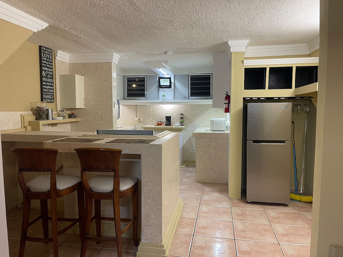 A spacious kitchen area features a large countertop with two wooden stools. Light-colored cabinetry and a stainless-steel refrigerator are visible, along with a modern stove and sink. Natural light is introduced through windows above the countertop, illuminating the tile flooring.