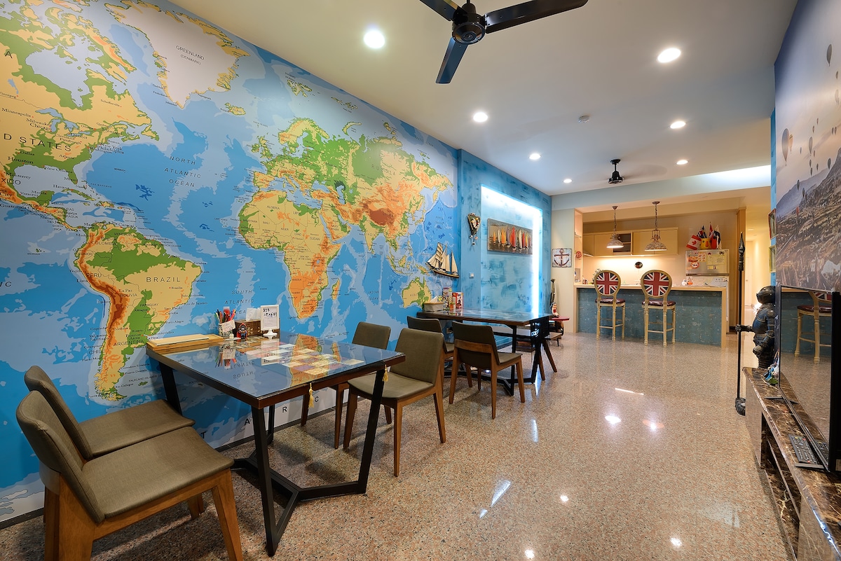 The communal area features a world map mural adorning the wall, accompanied by dark wood tables and light-colored chairs. Two bar-style seating areas with high stools are situated at the back, providing an inviting space for social interaction.