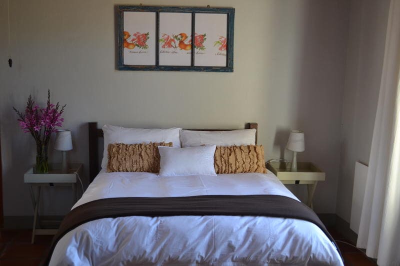 A spacious bed is centered in a softly lit room, adorned with decorative pillows and a neutral-colored blanket. Two small bedside tables with lamps are positioned on either side. Floral wall art adds a touch of color above the bed.