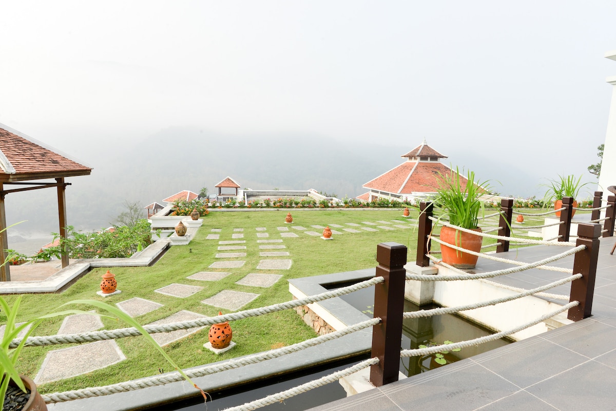 An expansive outdoor terrace is captured, featuring well-maintained grassy areas interspersed with stone pathways. Potted plants enhance the scenery, while fog envelops the distant mountains. The architecture includes charming buildings in the background, creating a serene atmosphere.