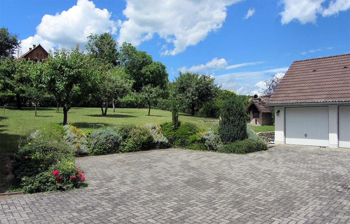 A spacious outdoor area is shown, featuring a stone-paved driveway and landscaped greenery. The scene is complemented by a variety of trees and flowering bushes. A house is visible in the background, and clouds drift in the blue sky above.