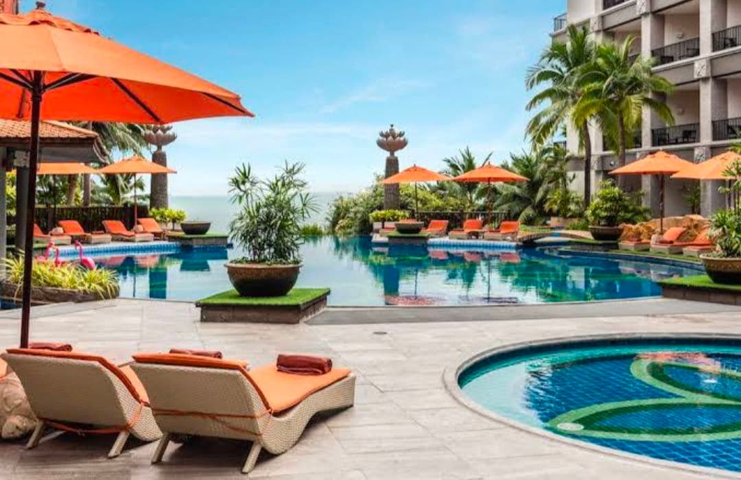A serene outdoor pool area features multiple lounge chairs under vibrant orange umbrellas. Lush greenery surrounds the pools, which reflect a clear sky. A jacuzzi is situated nearby, adding to the tranquil atmosphere.