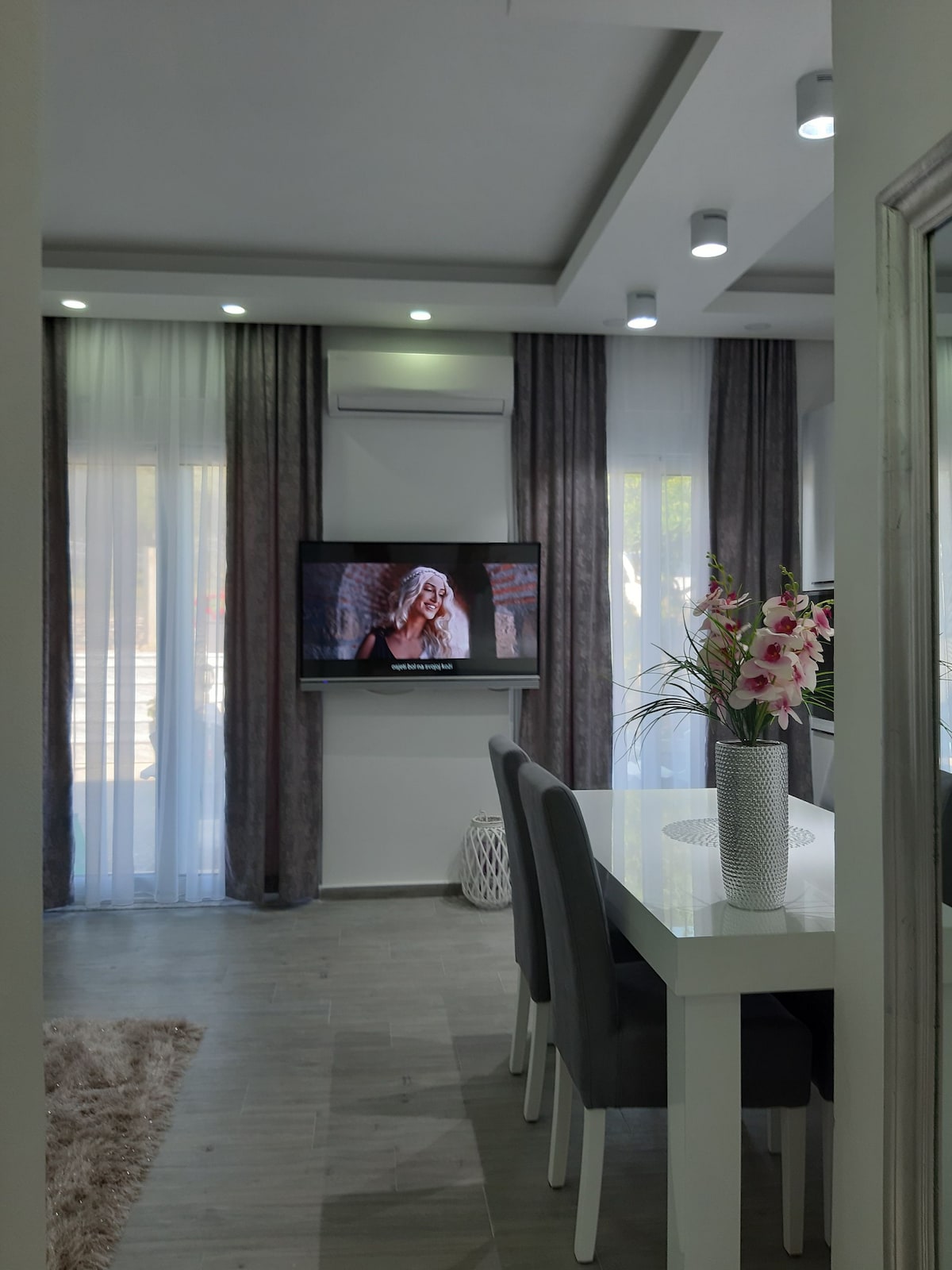 A dining area is seen with a white table and gray chairs, surrounded by large windows with sheer curtains allowing natural light to enter. A television mounted on the wall displays a program, while decorative flowers in a vase add a touch of color to the space.