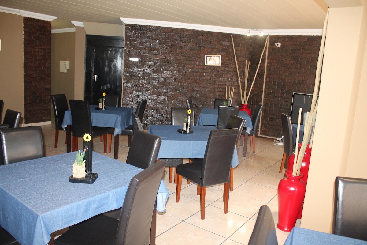 A dining area features several tables with blue tablecloths, accompanied by black leather chairs. The walls are finished in a rich, dark texture, and decorative elements, like potted plants and tall, slender vases, are positioned around the room.