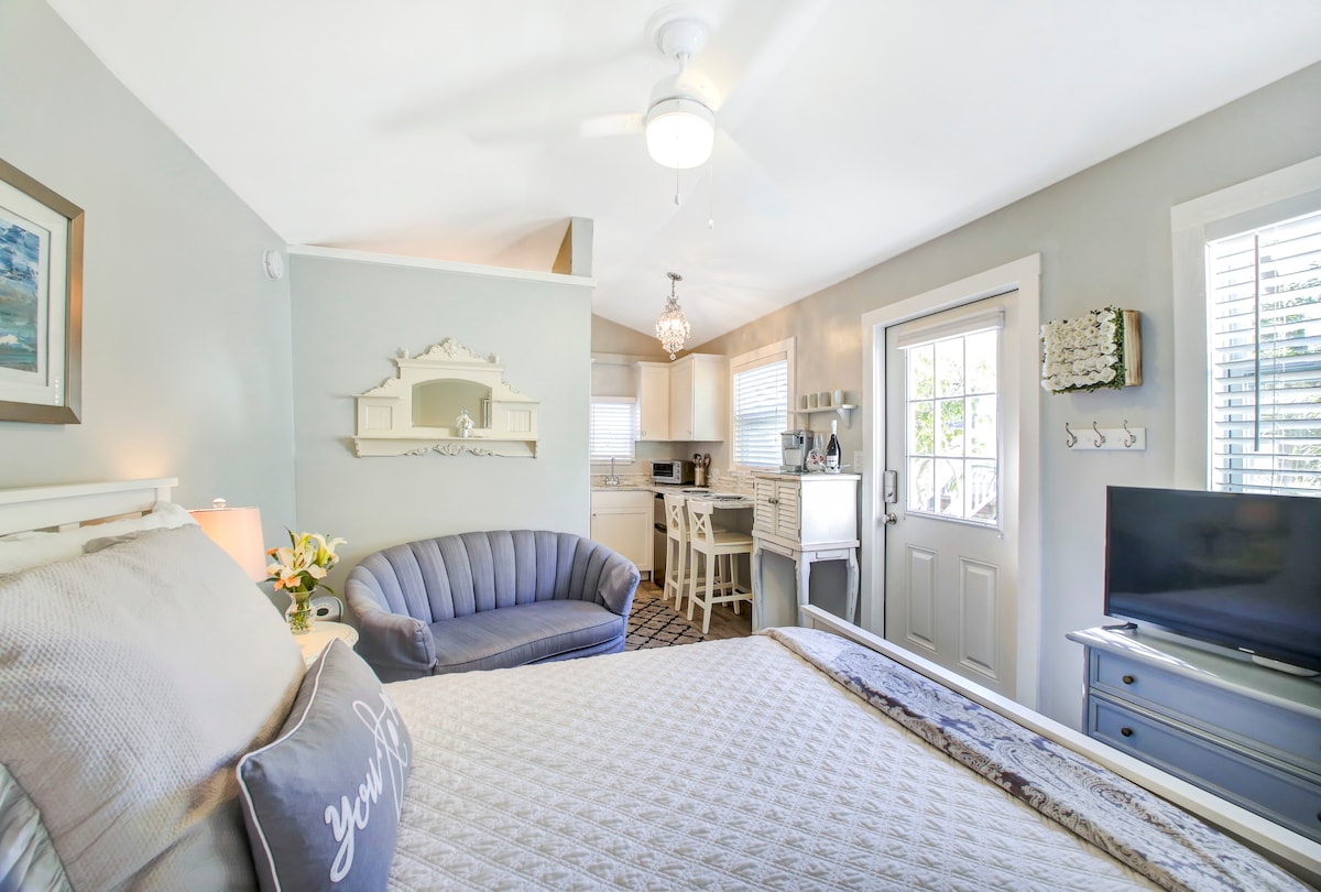 The studio features a queen bed with soft linens and a decorative pillow. A compact kitchen area with modern appliances is visible in the background. A cozy seating area is highlighted by a curved sofa, and natural light fills the space through large windows.