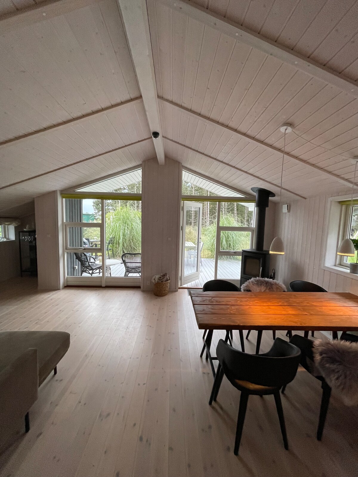 A bright communal area is presented with a large dining table surrounded by black chairs. Natural light floods in through expansive glass doors that lead to a deck, creating a harmonious connection with the outdoors. A wood-burning stove is visible, providing a cozy focal point.