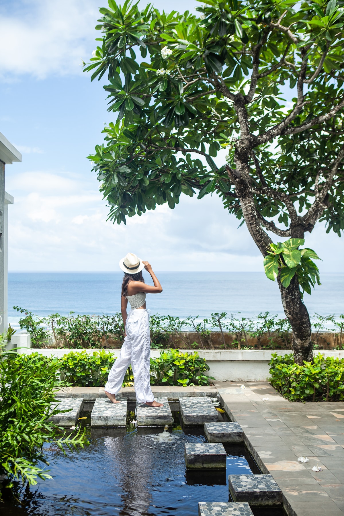 A person stands on stepping stones over a tranquil pool, overlooking the ocean. Lush greenery frames the view, enhancing the serene atmosphere. A tree with broad leaves extends overhead, adding to the natural setting. Soft clouds drift lightly across a bright blue sky.