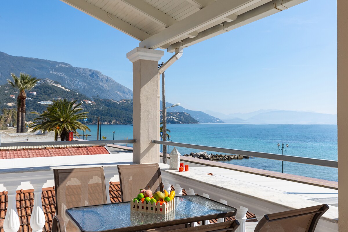 A spacious balcony is equipped with a glass dining table and four metal chairs, overlooking the Ionian Sea. Palm trees can be seen along the shoreline against a backdrop of distant mountains. A small basket of colorful fruit is placed centrally on the table.