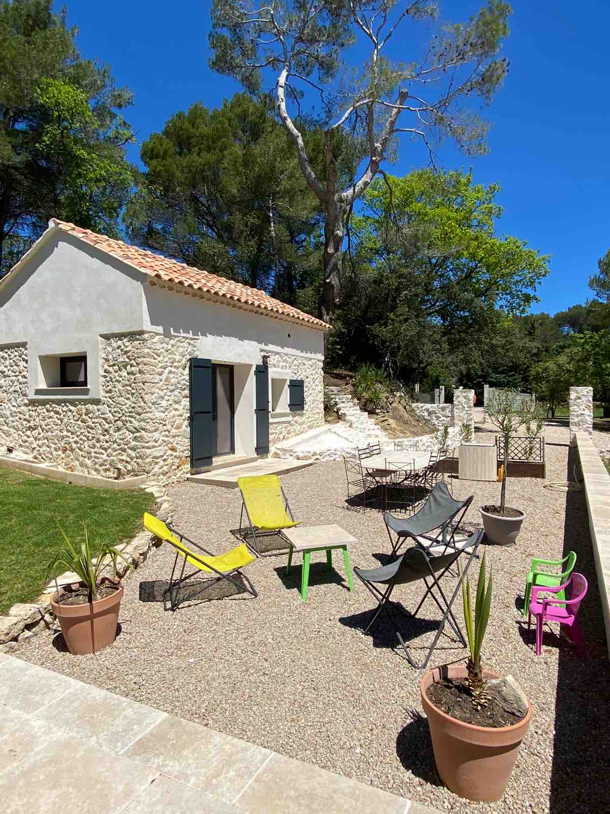A charming stone house is set against a backdrop of green trees, surrounded by a gravel area. Outdoor seating options include colorful chairs and potted plants, creating a welcoming space for relaxation. The pathway leads towards the entrance of the home, enhancing the serene outdoor environment.