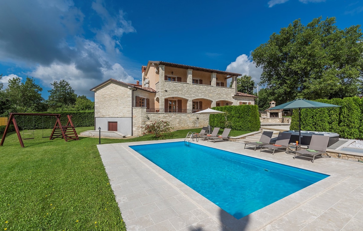 A two-story villa is set against a clear blue sky, surrounded by greenery. A swimming pool is positioned in front, accompanied by lounge chairs and umbrellas. A play structure and well-maintained lawn offer ample outdoor space for recreation. The house features a mix of stone and modern architecture.