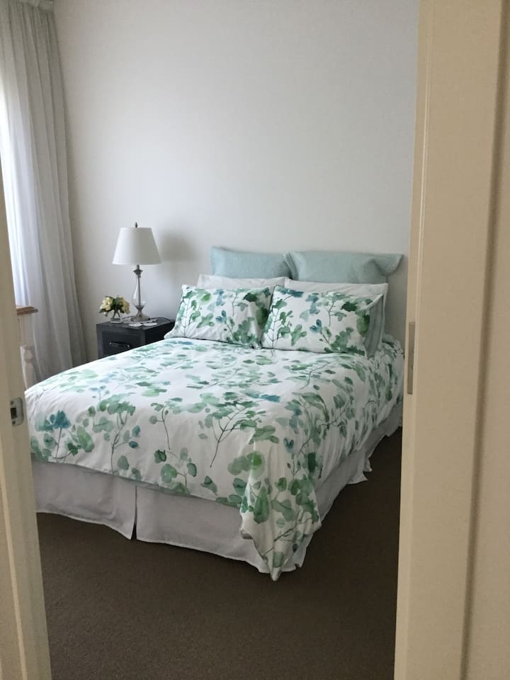 A small bedroom with a neatly made bed featuring green and white floral bedding. A bedside table holds a lamp and a small vase of flowers, with light curtains covering the window. The room has a soft, tranquil atmosphere with minimal decor.