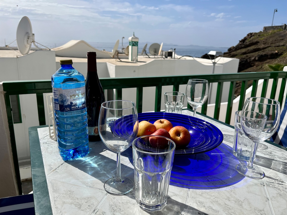 A table set on a terrace is adorned with a blue plate featuring fresh apples. Glasses and a water bottle sit nearby, complemented by a bottle of wine. The serene sea view is visible in the background, highlighting a peaceful outdoor dining space.