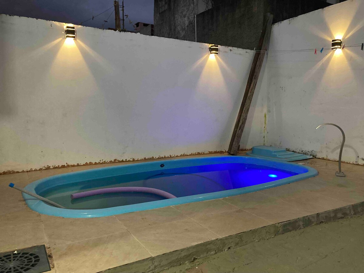 A softly lit pool area is surrounded by a clean white wall. The pool features gentle blue lighting, enhancing its inviting appearance. Two wall-mounted lamps provide warm illumination, and the surrounding paved area includes a small set of steps leading into the pool.