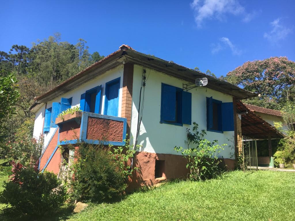 A charming countryside house is framed by lush greenery, showcasing blue shutters that contrast with the white façade. The structure features a staircase leading to the entrance, with vibrant plants enhancing the serene outdoor setting.