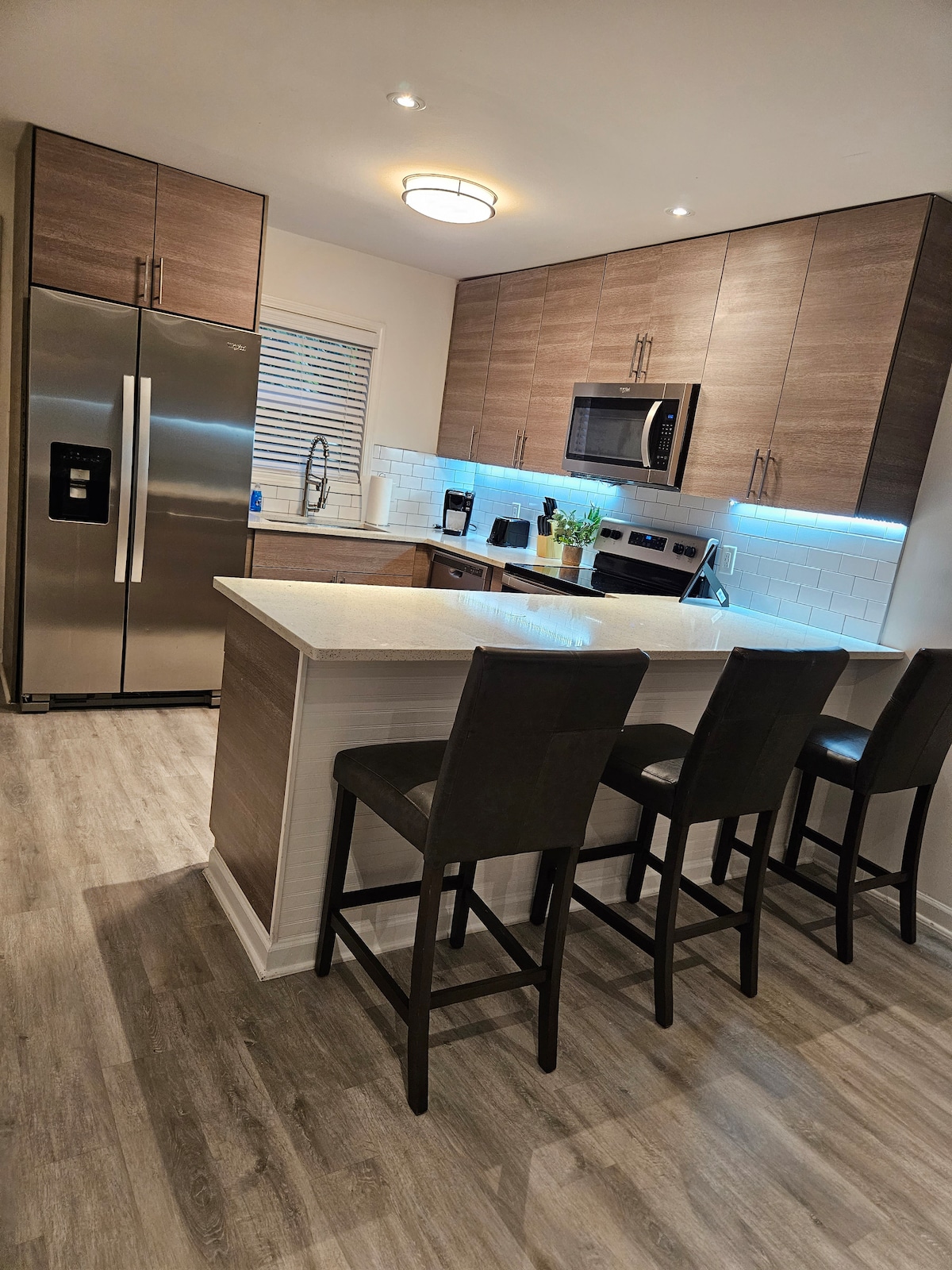 A modern kitchen features sleek cabinetry and stainless steel appliances. A spacious island with bar seating for three is highlighted by soft under-cabinet lighting. Natural light streams in through a window, enhancing the inviting atmosphere of the open layout.