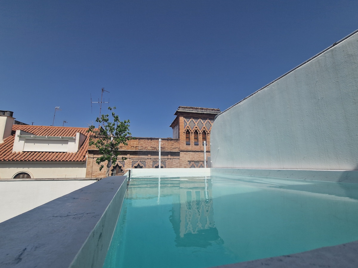 The image captures a rooftop pool, featuring clear turquoise water under a bright blue sky. Surrounding buildings are visible in the background, including architectural details such as arched windows. The pool's edge is framed by a white wall, enhancing the serene outdoor setting.