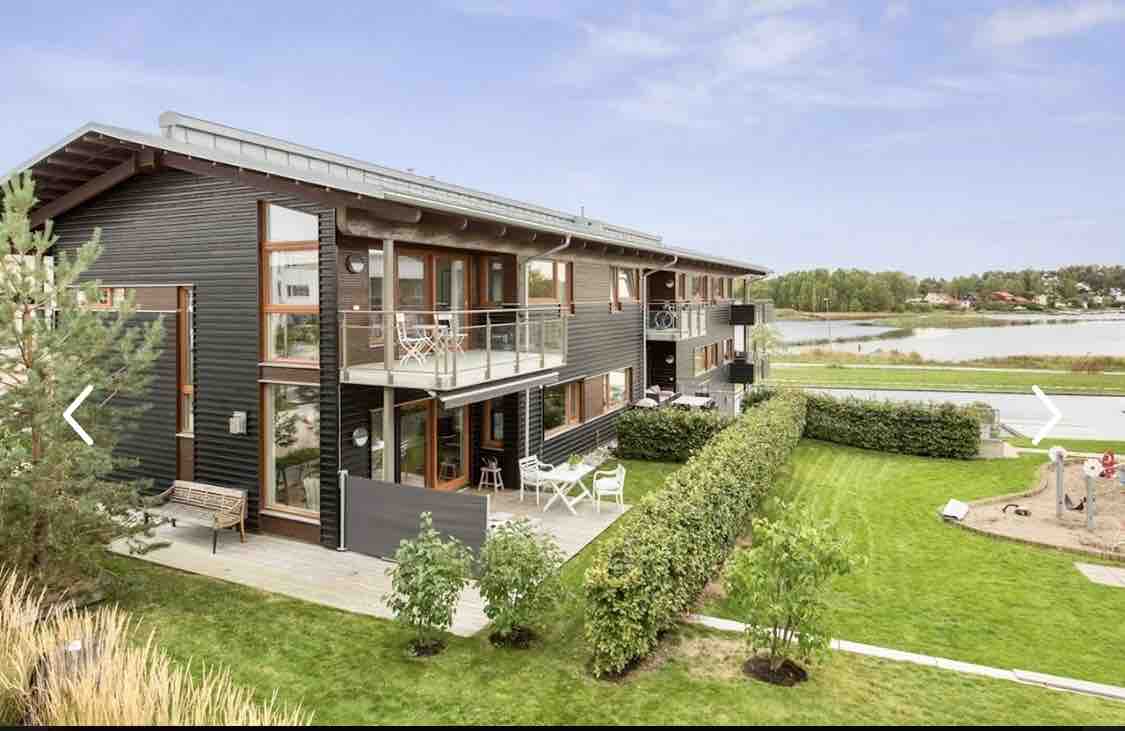 A modern wooden building features multiple balconies and large windows, allowing natural light to flood the interiors. Lush greenery surrounds the property, and the adjacent waterway is visible in the distance, providing a peaceful ambiance.
