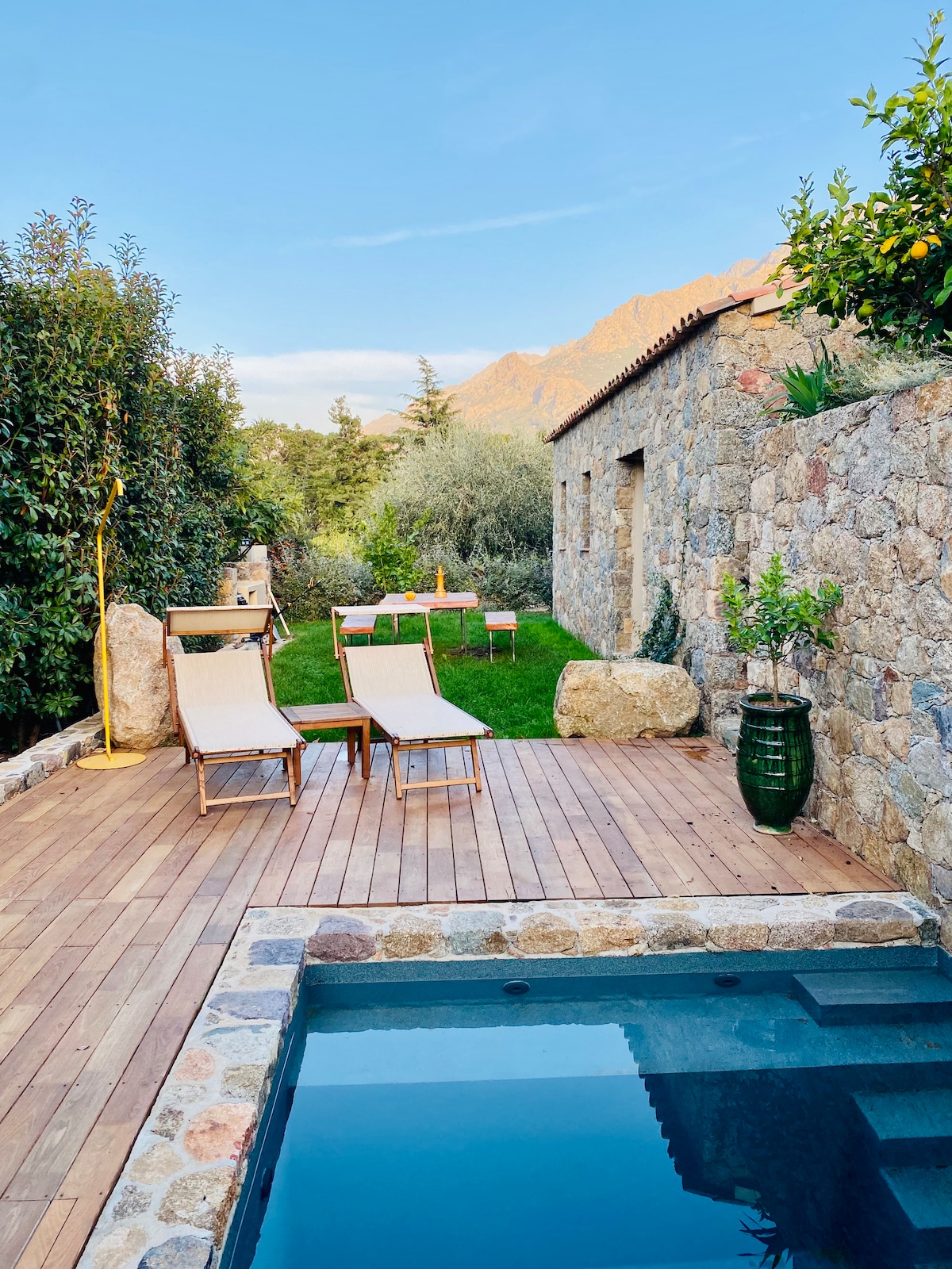 A serene outdoor space features a small heated pool surrounded by wooden decking. Two sun loungers are positioned nearby, offering a relaxing retreat. Lush greenery and natural stone walls create a tranquil atmosphere, with a wooden dining area visible in the background against the mountains.