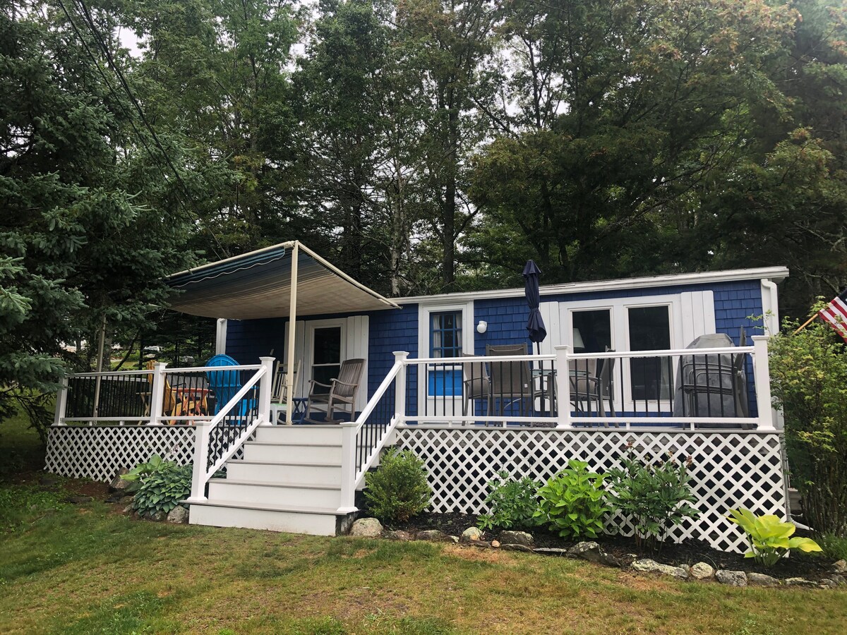A charming cottage exterior features blue siding and a white lattice deck. A covered porch extends along the front, furnished with seating and a dining area. Lush greenery surrounds the property, enhancing the peaceful outdoor setting.