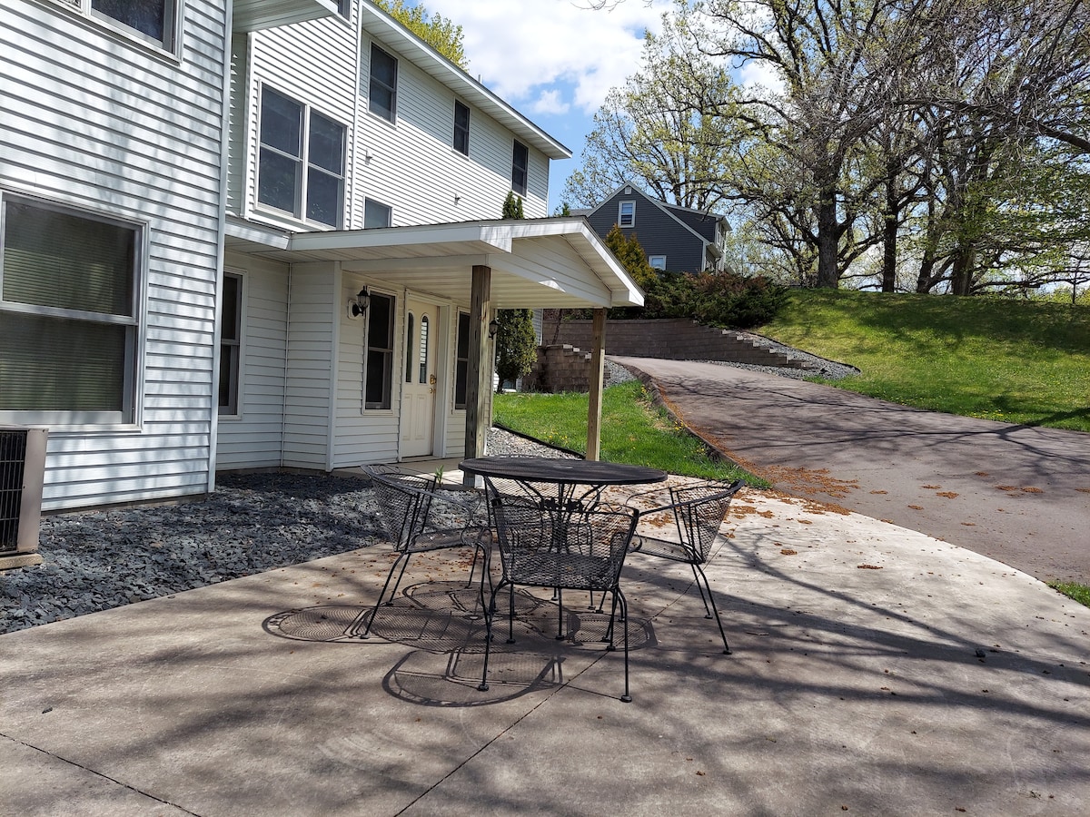 An outdoor patio area is visible, featuring a circular metal table surrounded by four matching chairs. The surface is paved, and the space is framed by a grassy area and trees in the background. An entrance to the building is located to the left.