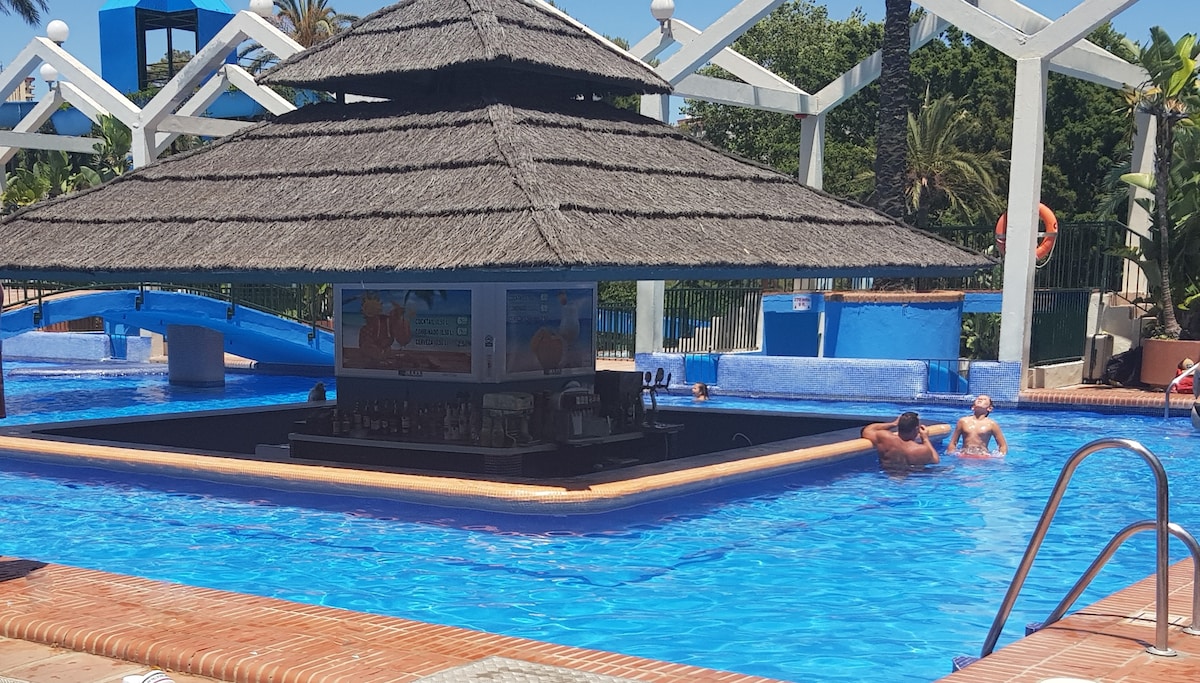 A swim-up bar is situated in the center of a large swimming pool, surrounded by palm trees and colorful slides. Guests are observed enjoying the water. The vibrant blue pool contrasts with the straw-roofed bar structure, creating a lively and inviting swimming area.