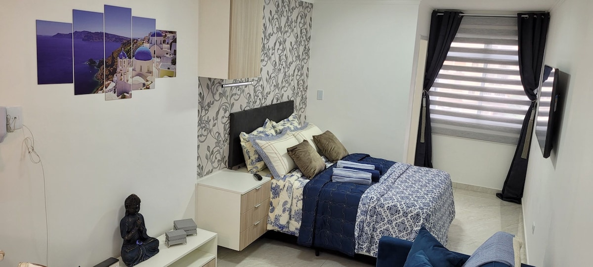 A cozy sleeping area features a spacious bed adorned with blue and white bedding. The wall displays art inspired by Santorini, and a decorative statue adds a touch of tranquility. Natural light filters through horizontal blinds, creating a calm atmosphere.