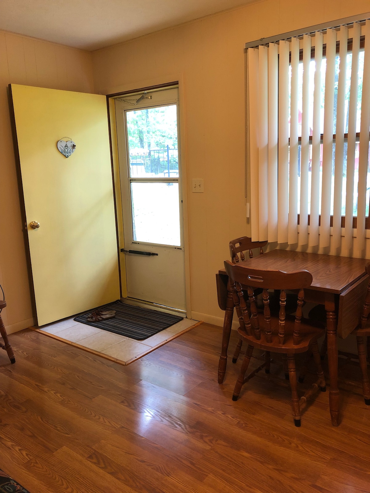 The entryway features a bright yellow door, welcoming guests into the cabin. A small wooden dining table with four chairs is positioned nearby, and vertical blinds offer a view to the outside. Light hardwood flooring provides a warm ambiance.