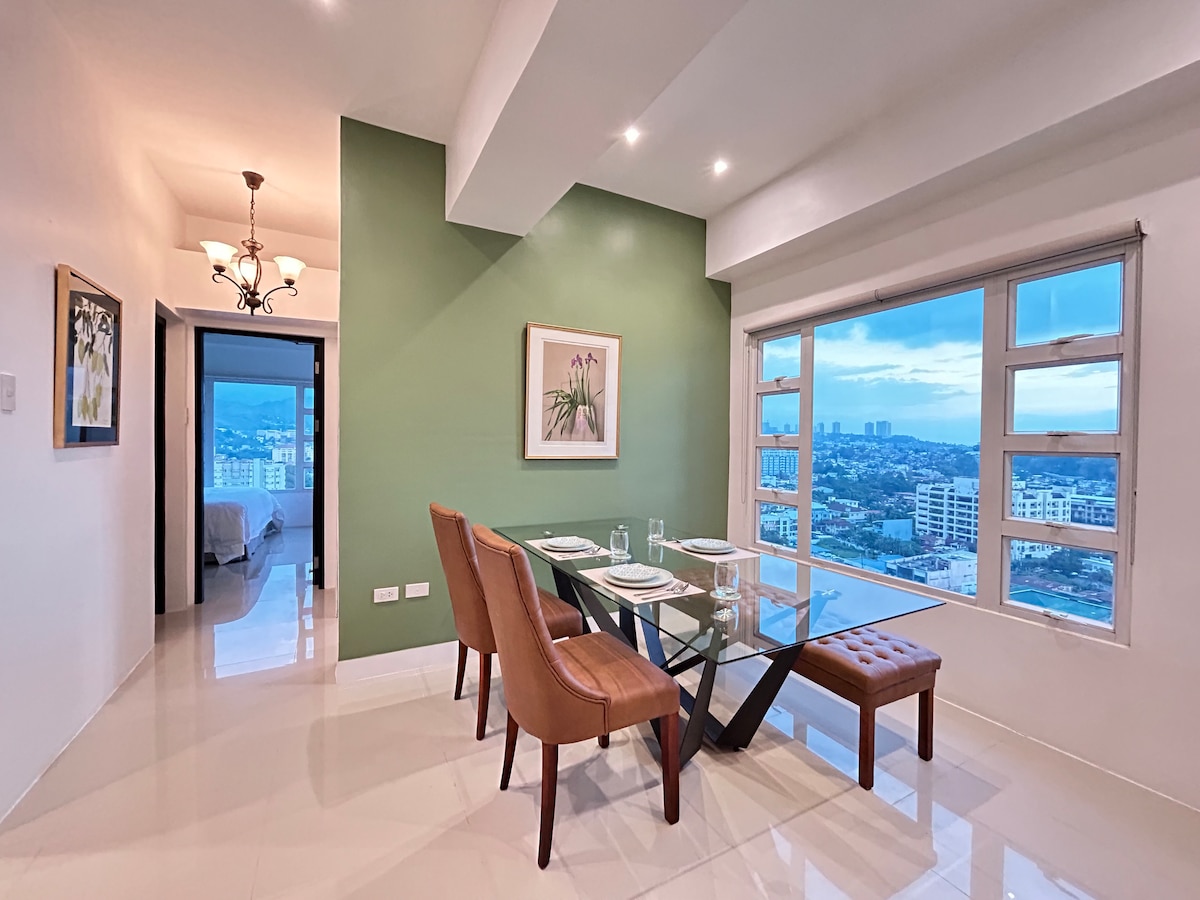 A dining area features a glass table accompanied by four upholstered chairs. A green accent wall adds color, while a framed artwork brings visual interest. Large windows provide a view of the cityscape, allowing natural light to brighten the space.