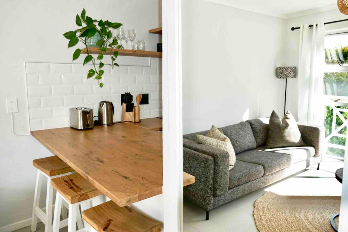 A modern kitchen area features a wooden countertop with seating for three and various small appliances. The adjacent living space has a comfortable gray sofa accented by cozy pillows, with natural light filtering through sheer white curtains.