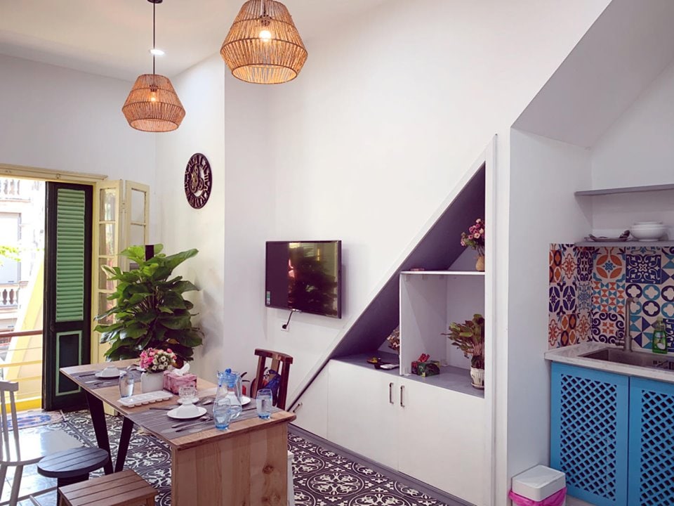 A bright dining area features a wooden table set for meals, accompanied by wooden chairs. The wall is adorned with a modern TV and decorative elements. Natural light filters through open windows with green shutters, enhancing the comfortable atmosphere accented by pendant lighting.