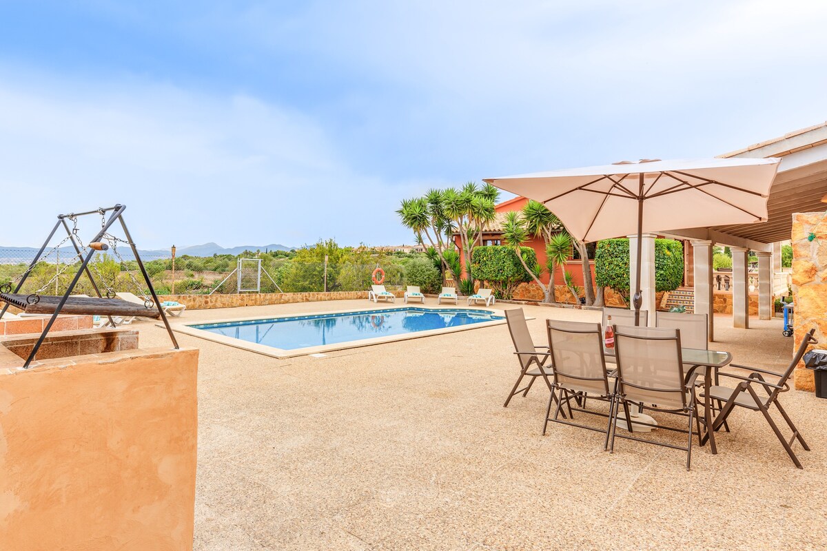 A spacious outdoor area features a saltwater pool and a large covered umbrella over a table with four chairs. The patio is surrounded by greenery and offers a scenic view of the distant sea and mountains.