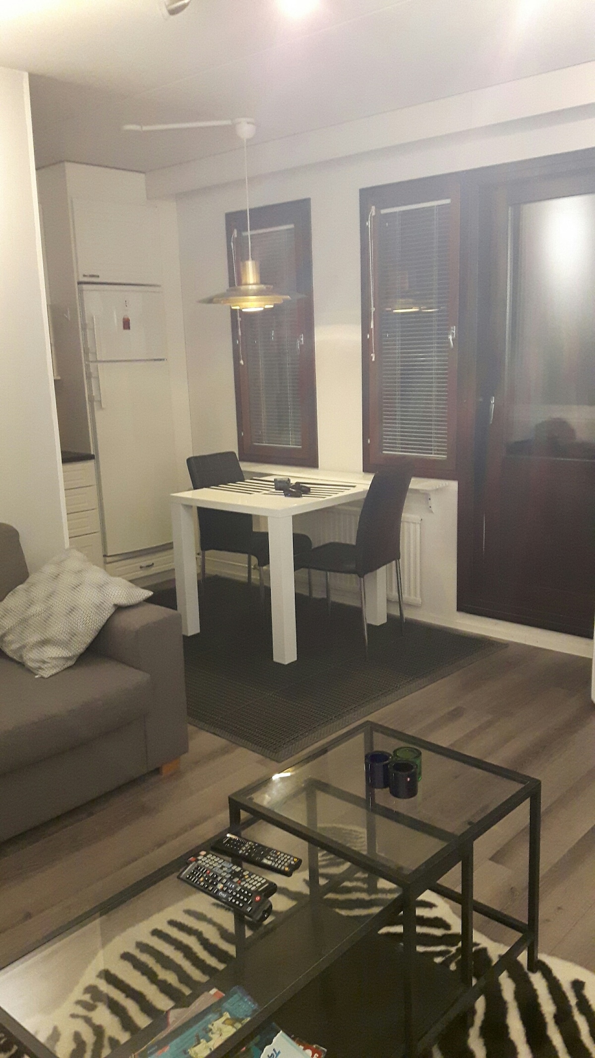 A comfortable living area is seen, featuring a gray sofa positioned near a glass coffee table. A dining table with two dark chairs is situated beside large windows covered by blinds. A modern kitchen is partially visible in the background.