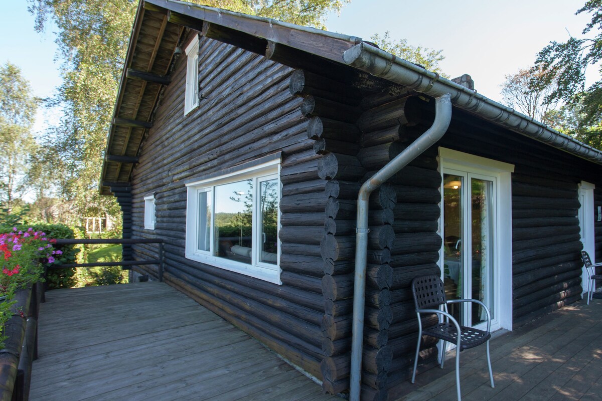 The exterior of the wooden chalet is displayed, featuring a large private terrace that invites outdoor relaxation. Two black chairs rest on the deck, and a large window offers views of the surrounding landscape. The structure's natural log design contributes to its rustic charm.