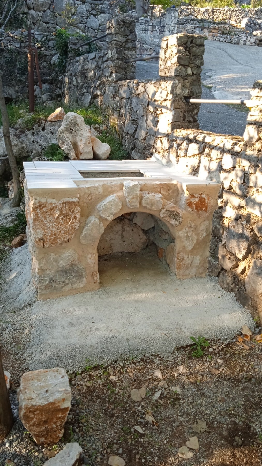 An outdoor stone well is situated amidst a natural setting, featuring a sturdy structure made of rough-hewn stones. The surrounding area is lined with additional stone features, enhancing the rustic charm of the landscape. The ground is mostly clear, highlighting the well's craftsmanship.
