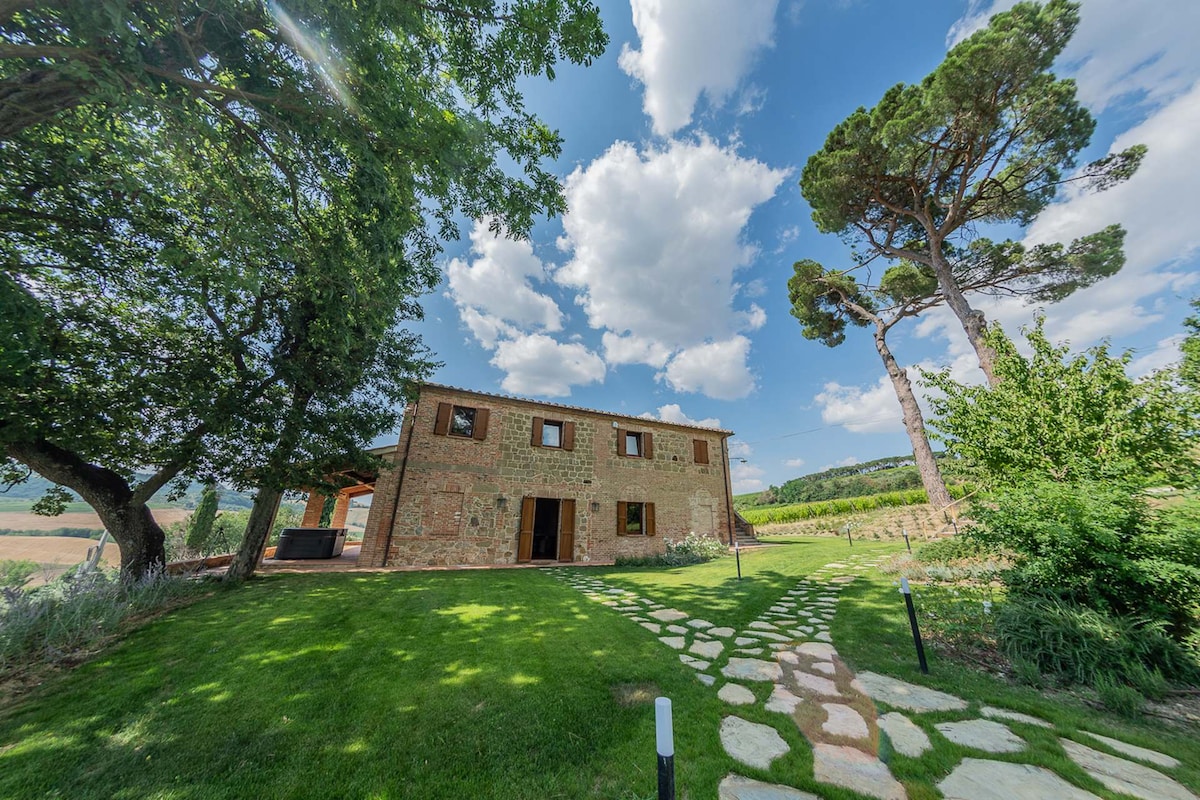 The exterior of the villa is framed by lush greenery and tall trees, with a stone pathway leading to the entrance. The structure features traditional architecture, characterized by stone walls and wooden accents, set against a backdrop of rolling hills and a blue sky with scattered clouds.