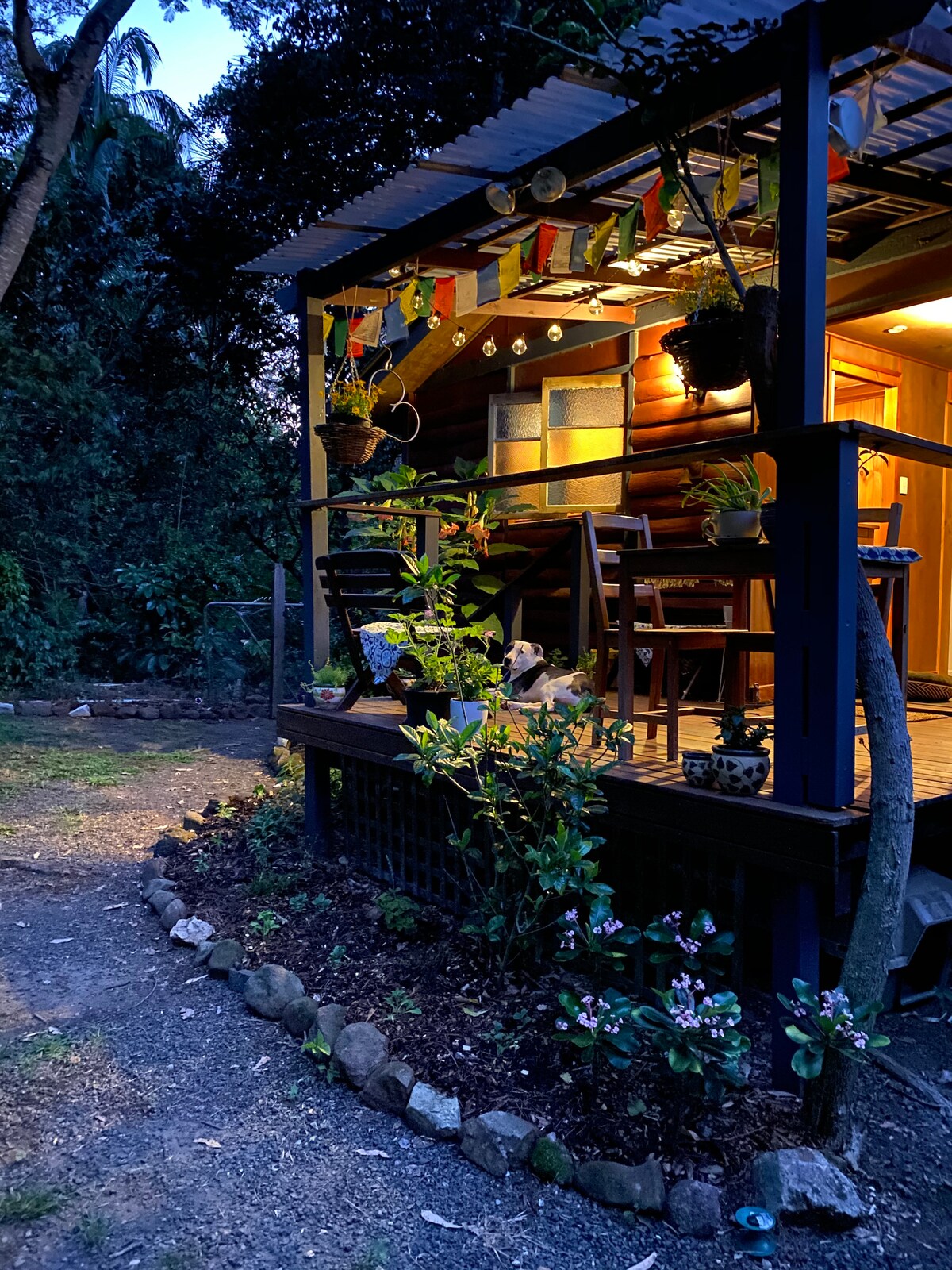 The log cabin exterior is softly illuminated at dusk, revealing wooden paneling and a welcoming covered balcony. Potted plants and colorful prayer flags adorn the space, while a gravel pathway leads towards the cabin, framed by a neat garden bed filled with greenery.