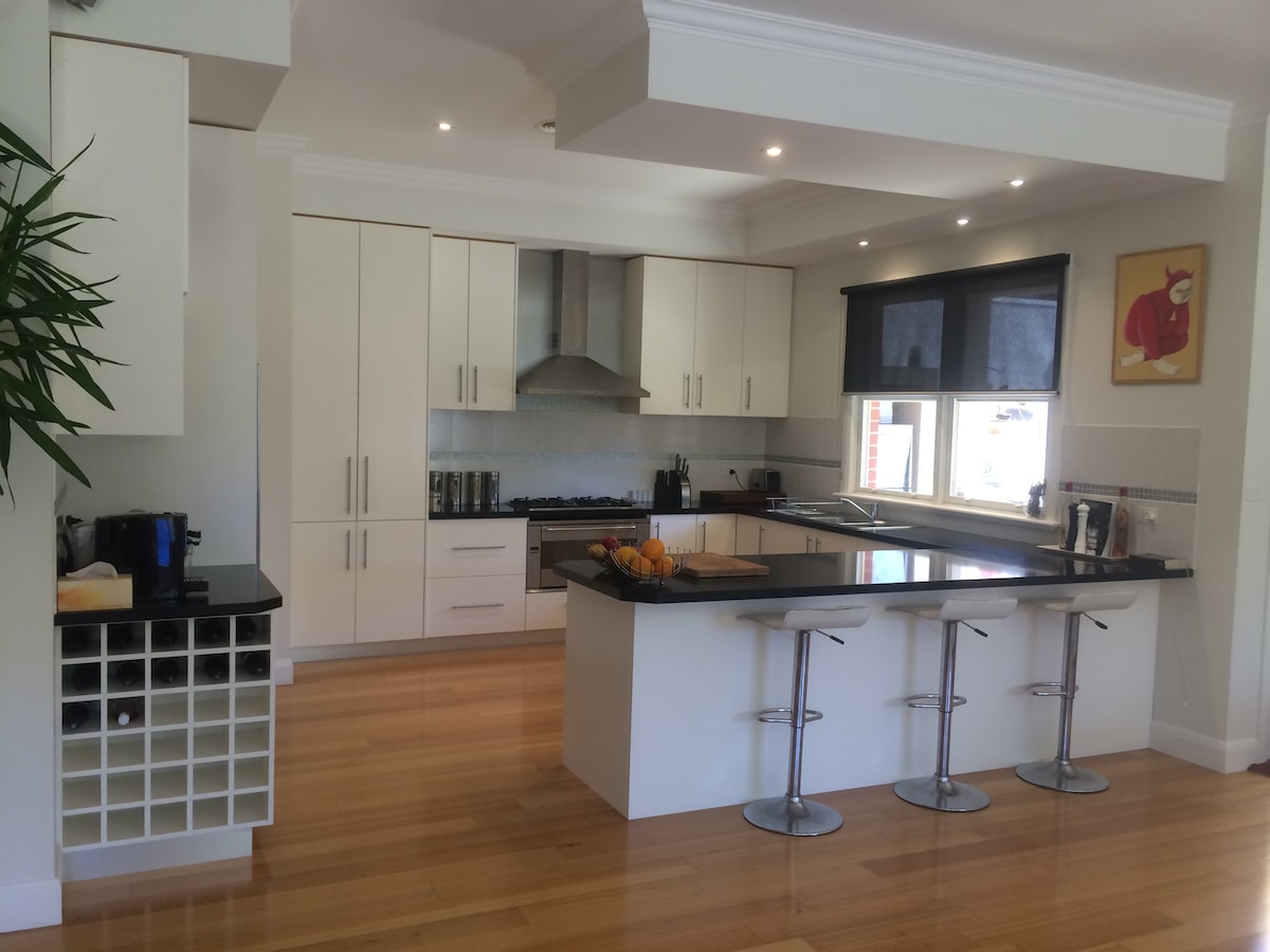 The modern kitchen features sleek cabinetry with ample storage and a spacious black countertop. Stainless steel appliances are visible alongside a breakfast bar with three stools. Natural light fills the area through a window, complemented by a fresh fruit bowl on the countertop.