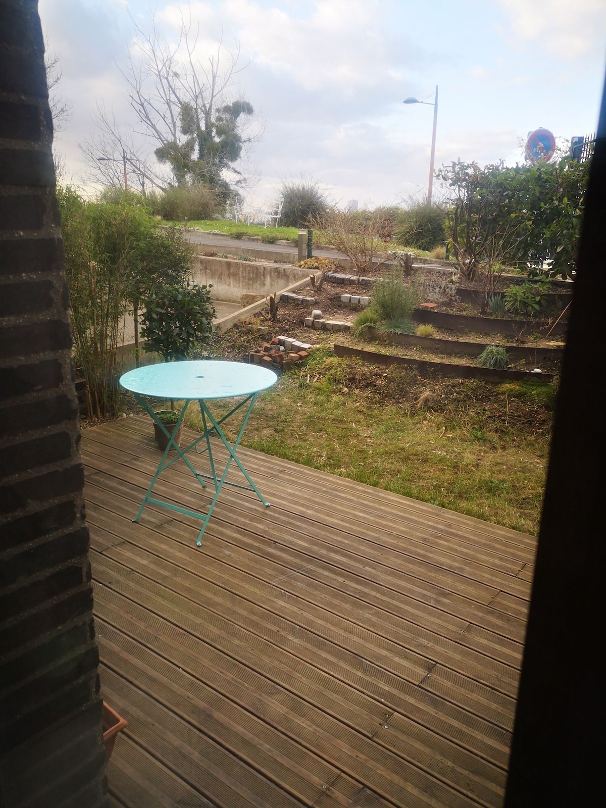 A small, round turquoise table is placed on a wooden deck. The table faces a garden area with various plants and trees, leading to a pathway lined with raised garden beds. Soft, natural light filters through the scene, highlighting the greenery.