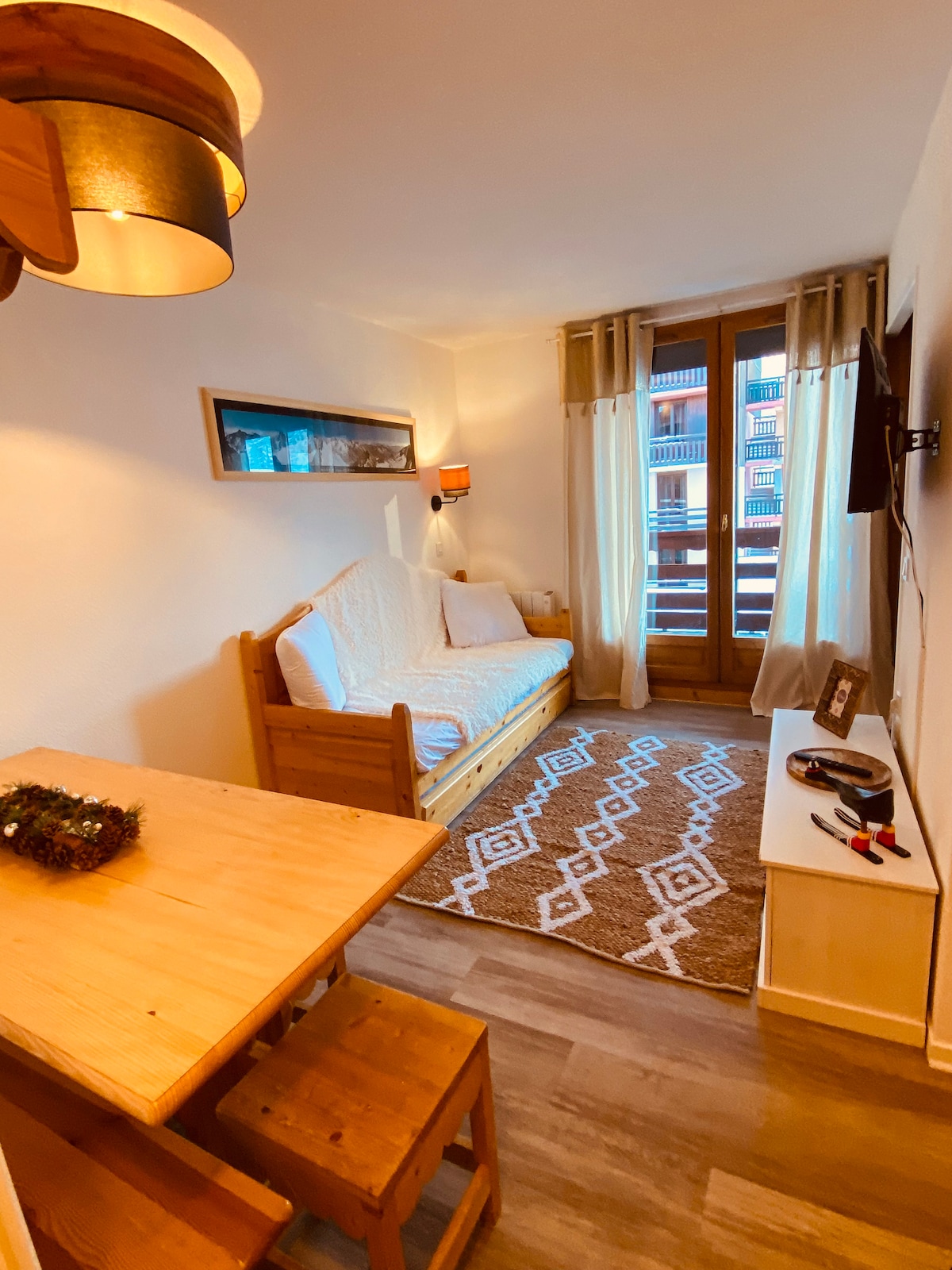 A cozy living area features a wooden table with two accompanying stools and a compact sofa bed. Natural light filters through the balcony doors, illuminating a patterned area rug. A wall-mounted flat-screen TV and a small decorative shelf add functionality to the space.