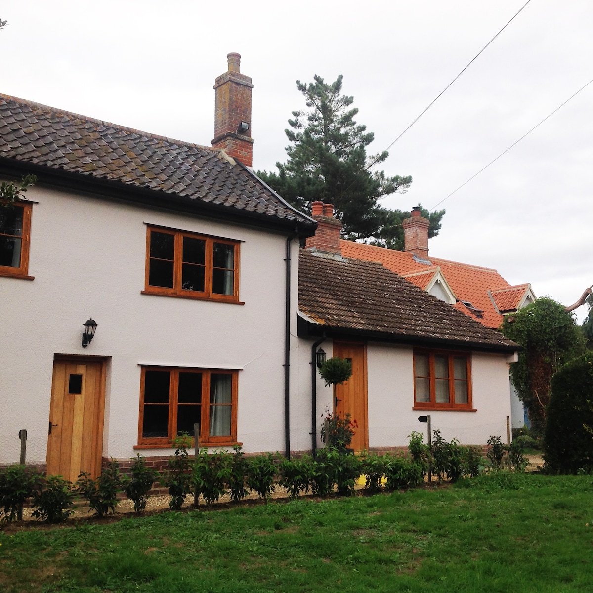 The exterior of a charming semi-detached cottage is presented, featuring a light-colored facade with wooden accents. A well-maintained garden with low bushes surrounds the property, and traditional chimneys rise from the roof. The peaceful setting is complemented by a gravel path leading to the entrance.