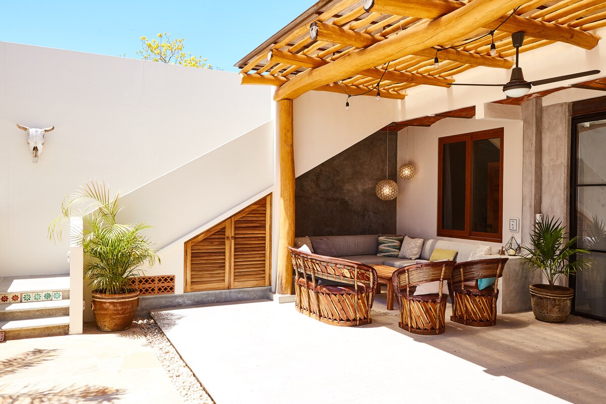A shaded patio features a built-in L-shaped couch adorned with soft cushions, surrounded by woven chairs. Decorative pendant lights hang above, while plants add a touch of greenery. The area is framed by light-colored walls and offers a cozy spot for relaxation.
