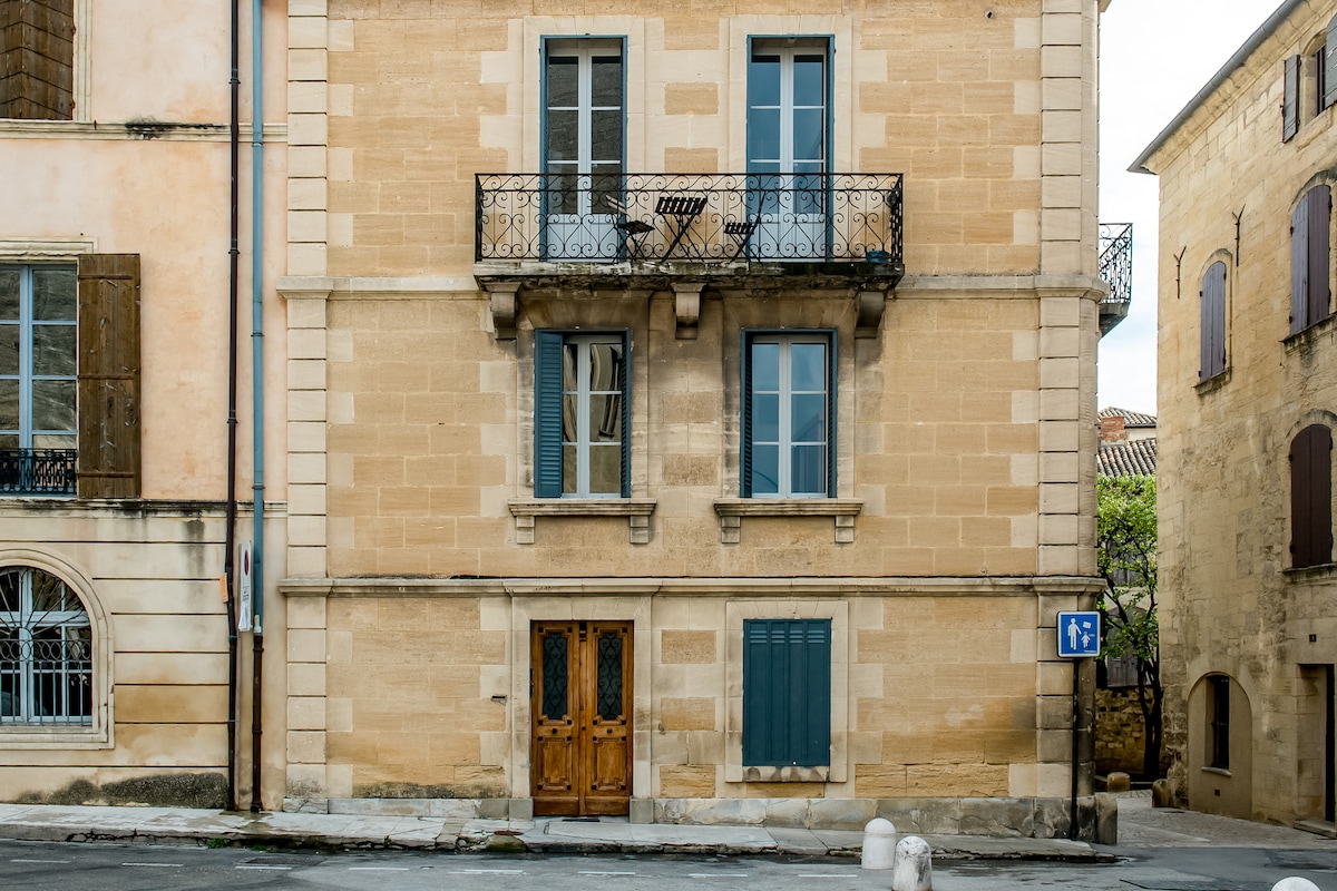 The exterior of a historic building is showcased, featuring light-colored stone walls and large windows with green shutters. A balcony adorned with an iron railing is positioned above the entrance, which is framed by wooden double doors. A street sign is visible to the right.