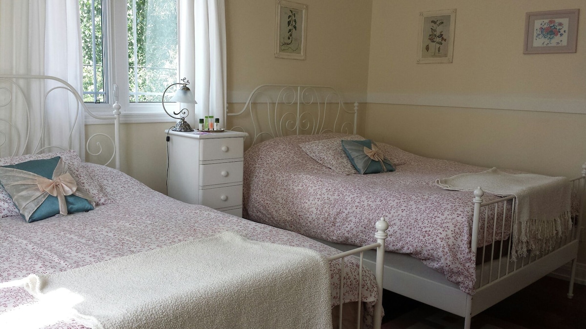 A bright room features two queen size beds with soft floral bedding. A white nightstand is placed between the beds, holding personal items. Natural light fills the space through a nearby window, with soft curtains allowing for a welcoming ambiance.