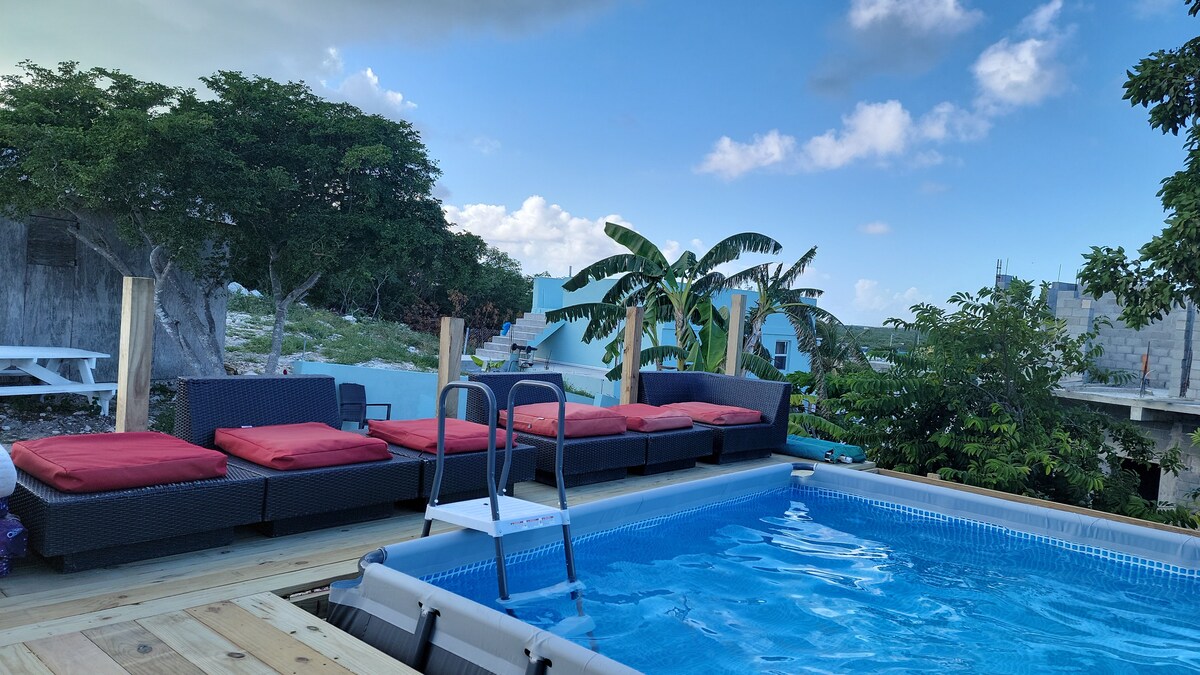 A private outdoor lounging area features several dark wicker lounge chairs with red cushions positioned near a sparkling blue pool. Lush greenery surrounds the space, creating a calm atmosphere. The pool includes a ladder for easy access and is bordered by a wooden deck.